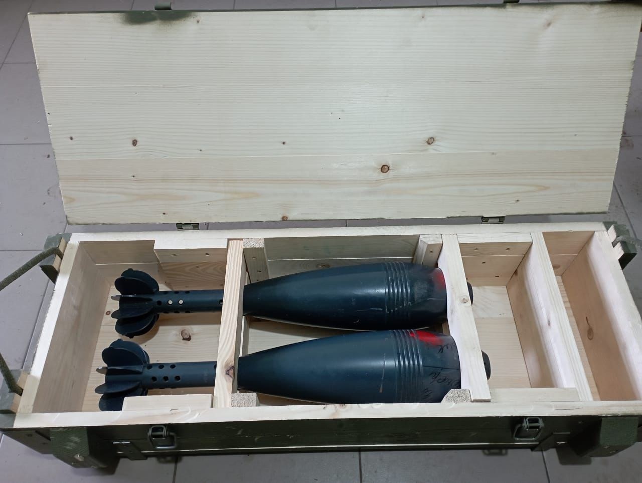 Two ammunition-shaped items supported inside a green wooden crate; live or inert status is not established from the photograph